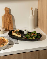 A kitchen counter with a black plate holding a slice of quiche, a green salad, and a small cup. Nearby are a white bottle with a cork, a wooden cutting board, garlic, and a whole quiche in a white dish